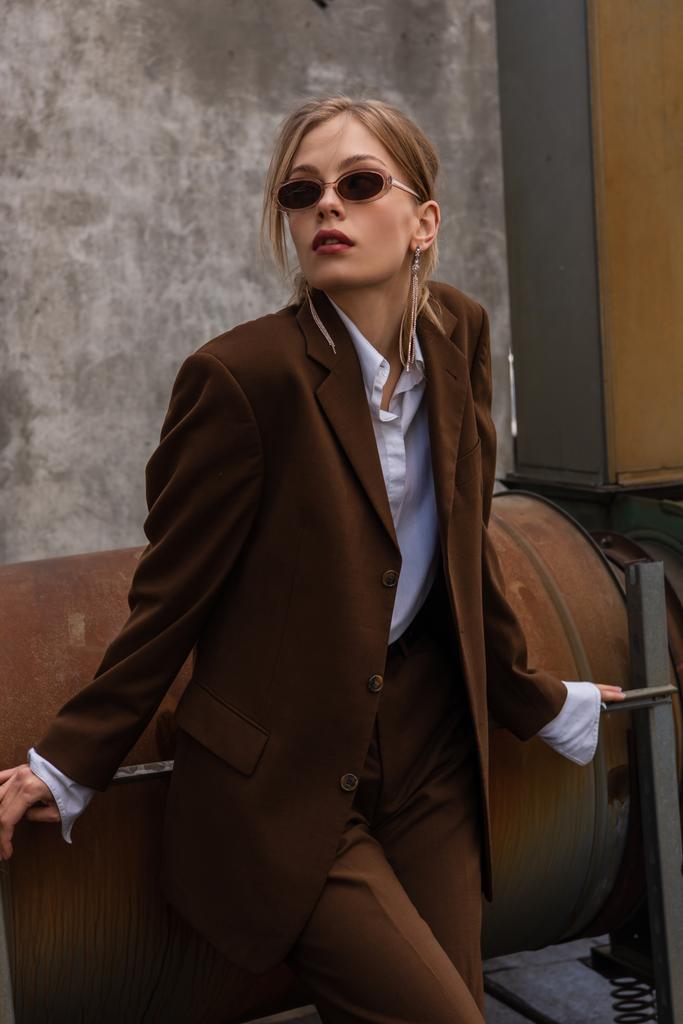mladá blondýna modelka v slunečních brýlích a módní hnědý oblek pózuje na střeše  - Fotografie, Obrázek