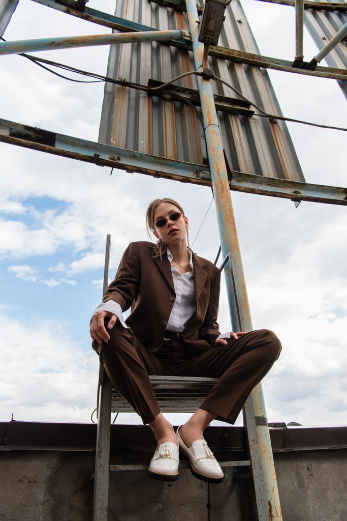 屋上の金属製の階段に座ってサングラスと茶色のスーツの若い女性の低角度ビュー - 写真・画像