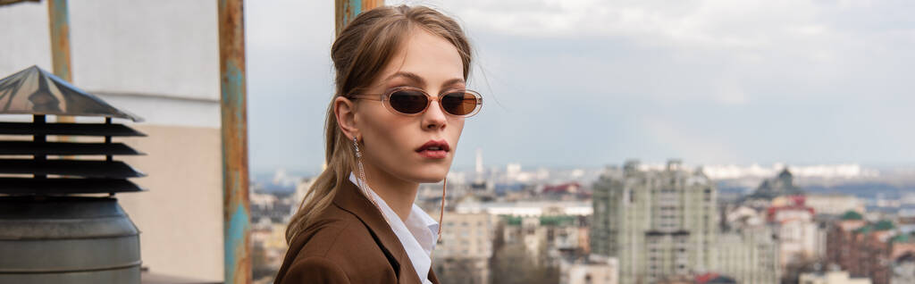 modelo joven en gafas de sol elegantes posando en la azotea con paisaje urbano sobre fondo borroso, pancarta - Foto, imagen