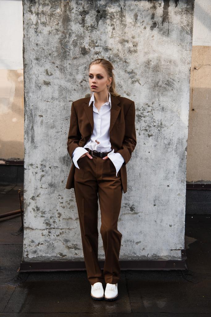 full length of young woman in stylish suit posing with hands in pockets near concrete wall - Photo, Image