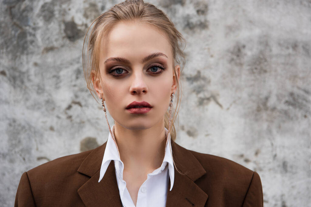 giovane donna in orecchini alla moda e blazer posa vicino muro di cemento - Foto, immagini