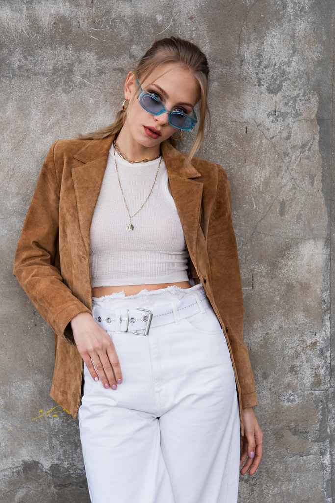 модна молода модель в блакитних сонцезахисних окулярах і стильному вбранні позує біля бетонної стіни
 - Фото, зображення
