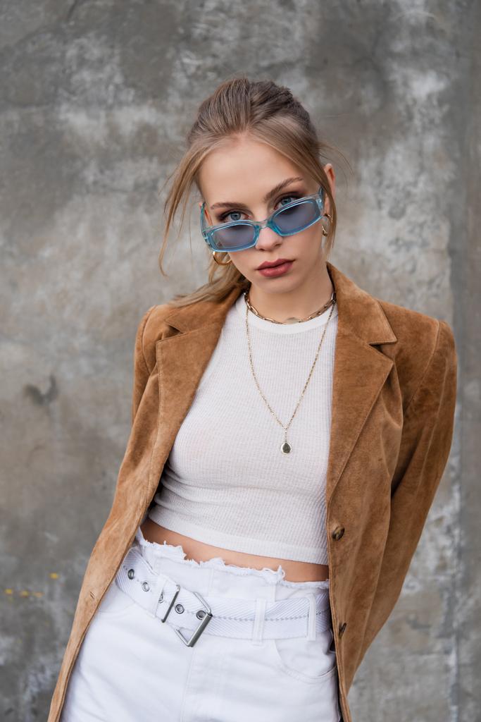 model v modrých slunečních brýlích a stylové oblečení pózující v blízkosti šedé betonové stěny - Fotografie, Obrázek