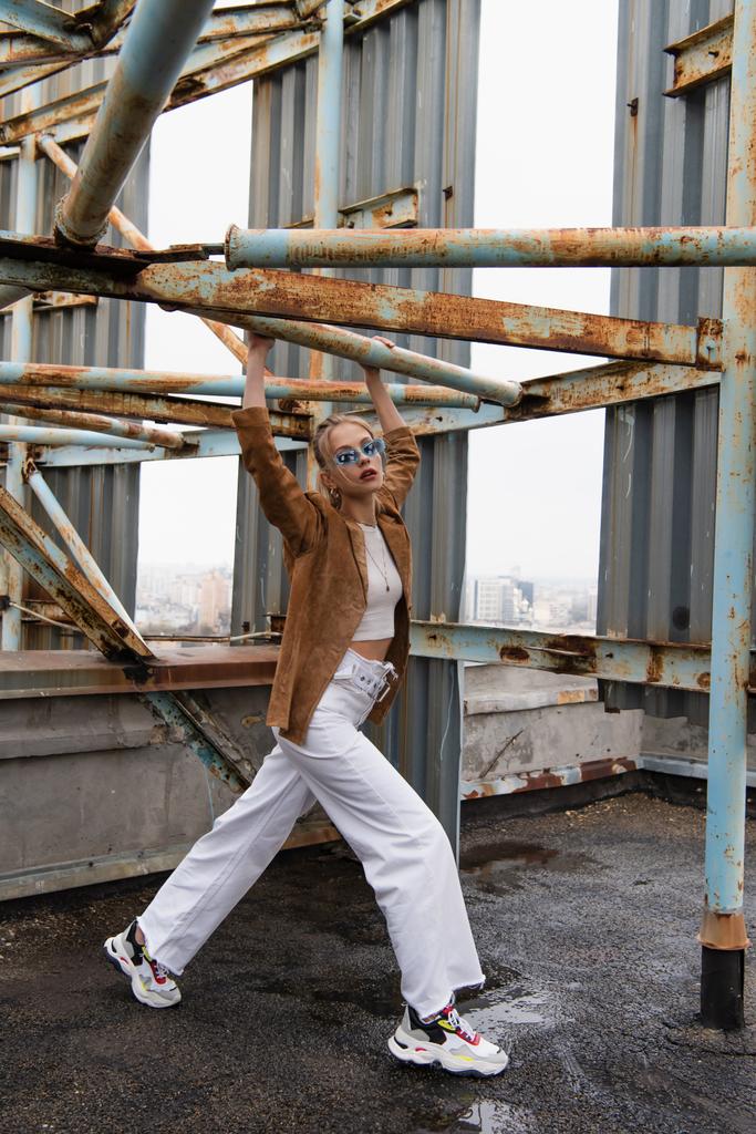 full length of trendy young model in blue sunglasses posing near rusty construction on rooftop - Photo, Image