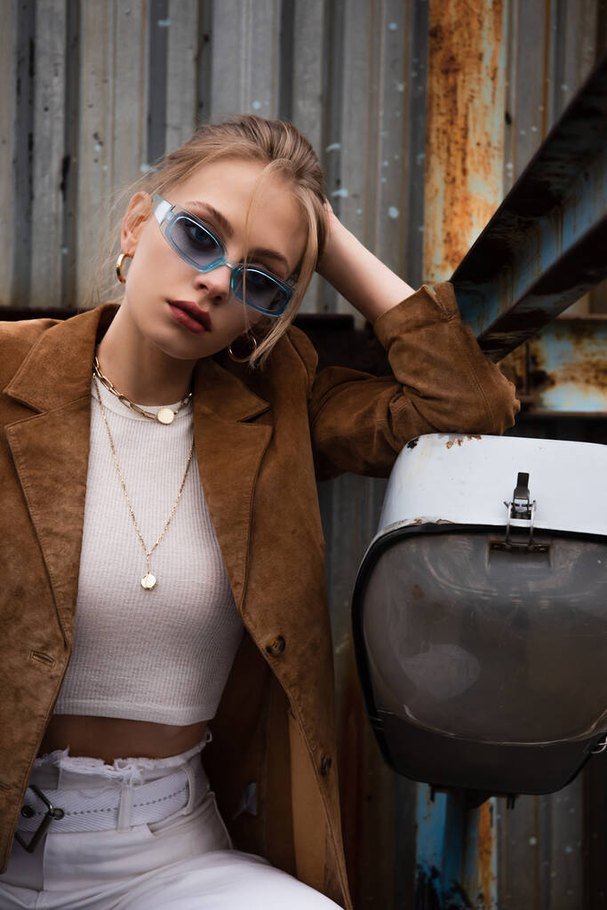 blonde woman in blue sunglasses and suede jacket sitting while posing on rusty rooftop  - Photo, Image