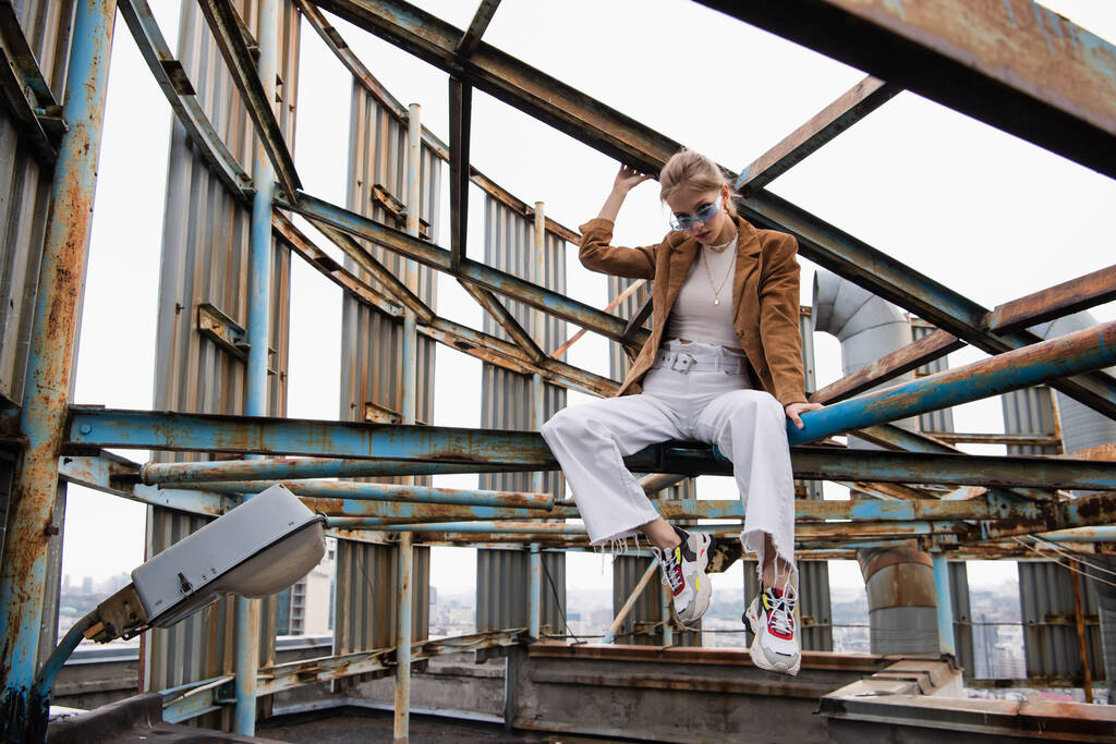 young woman in blue sunglasses and trendy outfit sitting on rusty construction of rooftop  - Photo, Image