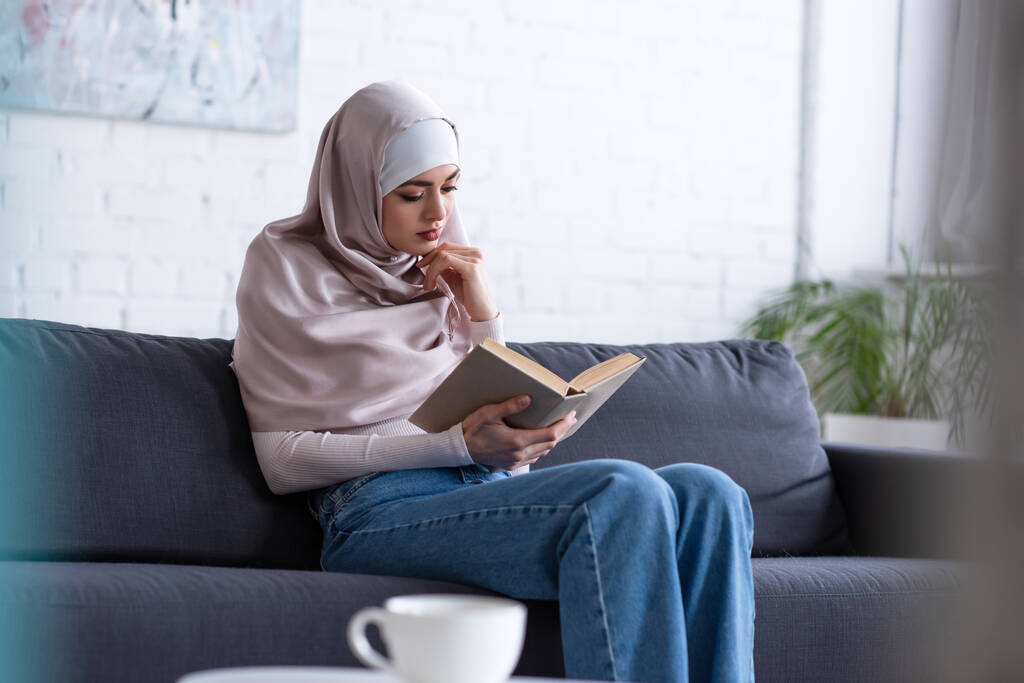 melko muslimi nainen lukee kirjaa sohvalla lähellä hämärtynyt kuppi teetä - Valokuva, kuva