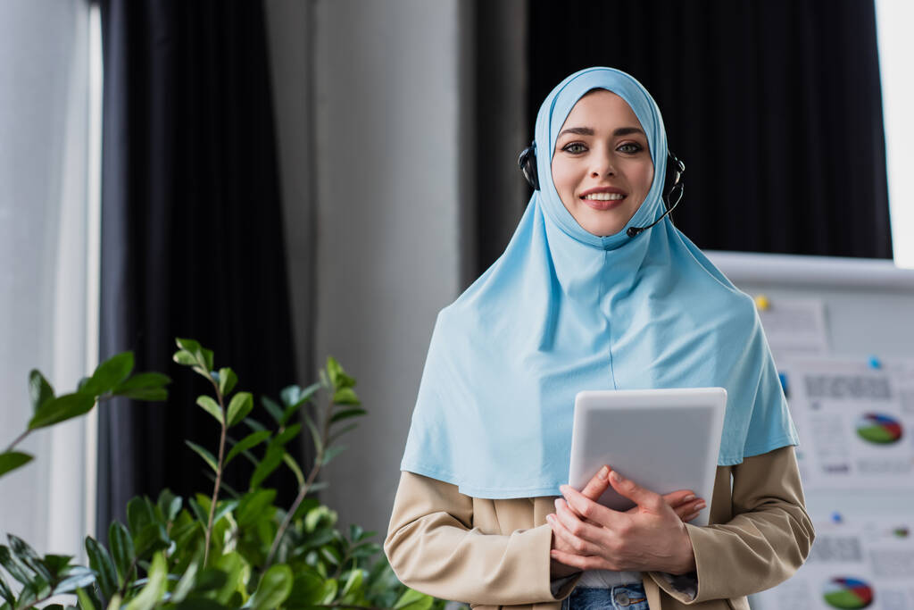 радісна арабська жінка в хіджабі і гарнітурі посміхається на камеру, тримаючи цифровий планшет
 - Фото, зображення