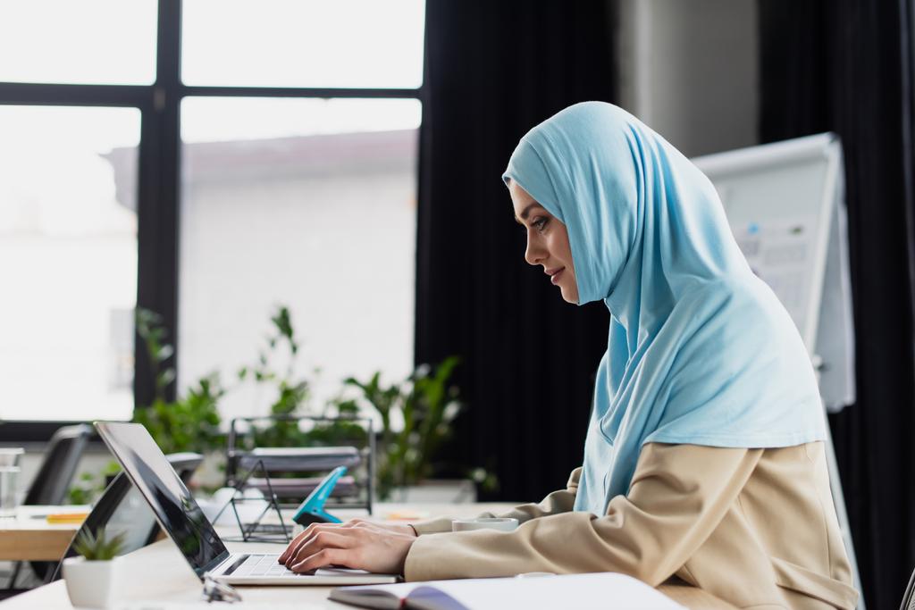 πλαϊνή άποψη της νεαρής αραβικής επιχειρηματία σε hijab δακτυλογράφηση σε φορητό υπολογιστή στο γραφείο - Φωτογραφία, εικόνα