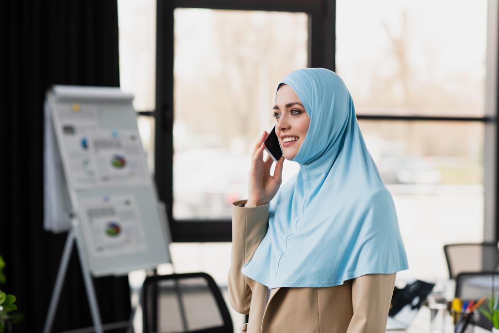 sonriente musulmana mujer de negocios hablando en el teléfono inteligente cerca de rotafolio borrosa - Foto, Imagen