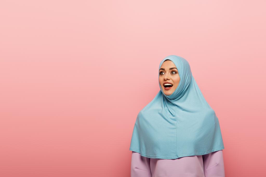 emocionada mujer musulmana en hijab azul mirando hacia otro lado aislado en rosa - Foto, Imagen