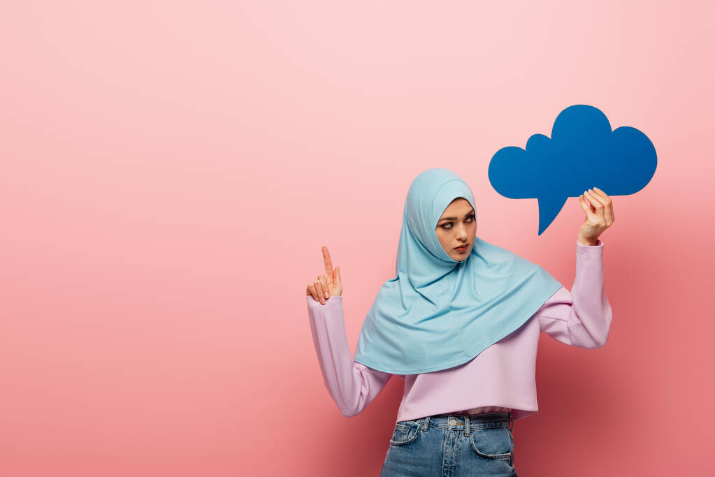 mujer musulmana con burbuja de pensamiento azul apuntando con el dedo sobre fondo rosa - Foto, imagen