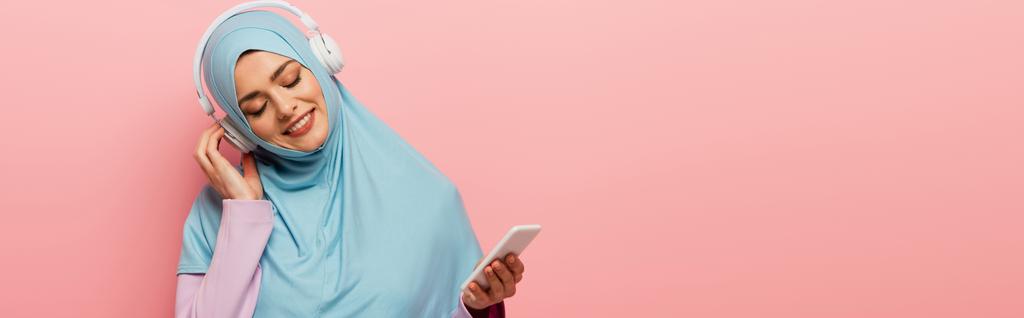 femme arabe positive en hijab bleu écoutant de la musique dans des écouteurs sans fil isolés sur rose, bannière - Photo, image