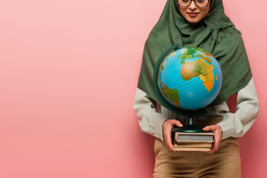 bijgesneden beeld van moslim leraar houden wereldbol en leerboeken over roze achtergrond - Foto, afbeelding