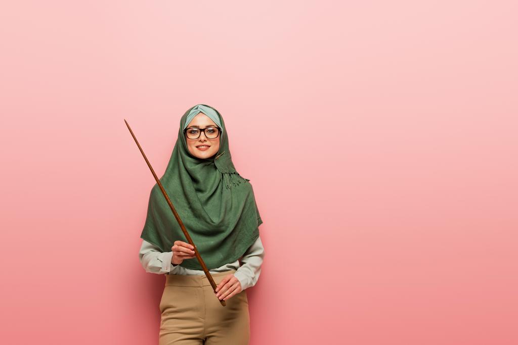 Genç Müslüman öğretmen kameraya gülümserken pembe arka planda işaret çubuğu tutuyor. - Fotoğraf, Görsel