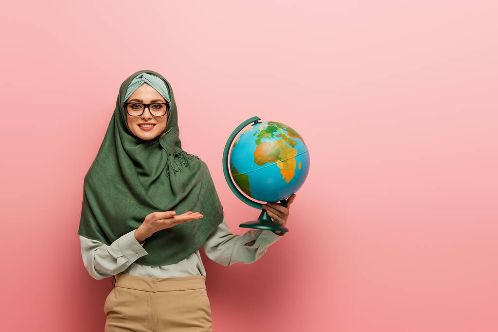 Yeşil tesettürlü gülümseyen Müslüman öğretmen pembe arka plandaki küreyi işaret ediyor. - Fotoğraf, Görsel