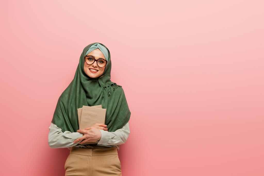 pretty muslim teacher with textbooks smiling at camera on pink background - Photo, Image