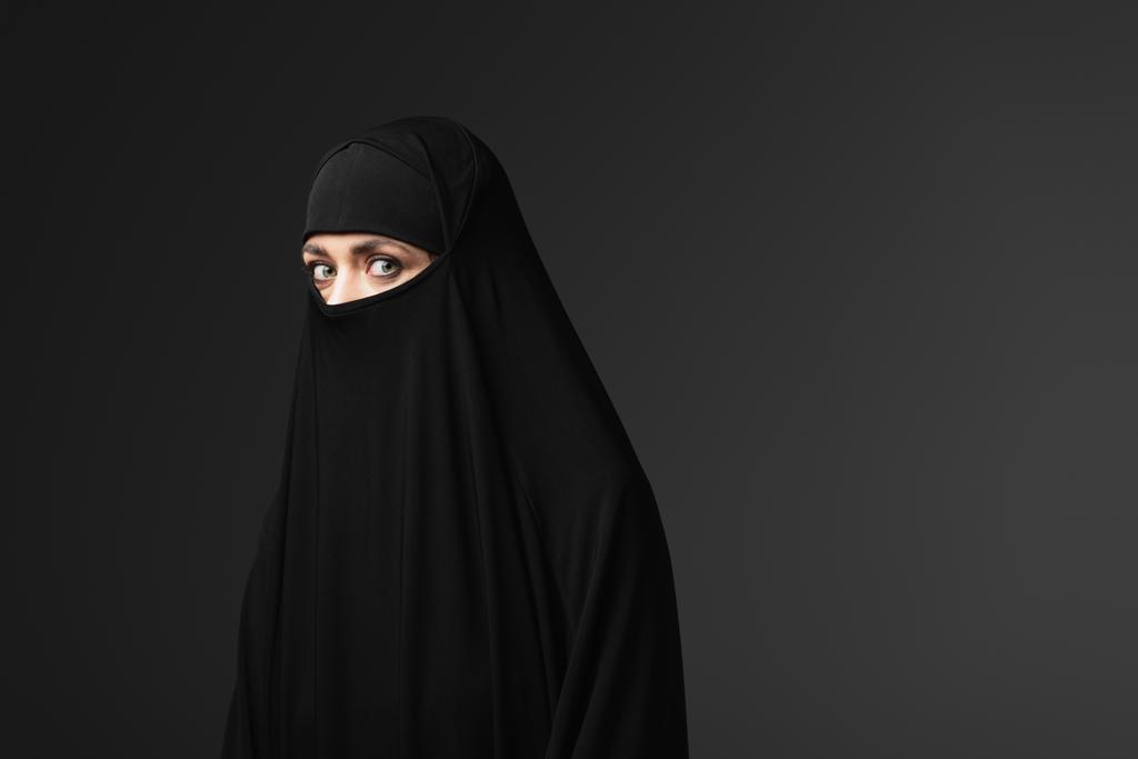 junge muslimische Frau in schwarzer traditioneller Kleidung blickt vereinzelt in die Kamera auf schwarz - Foto, Bild