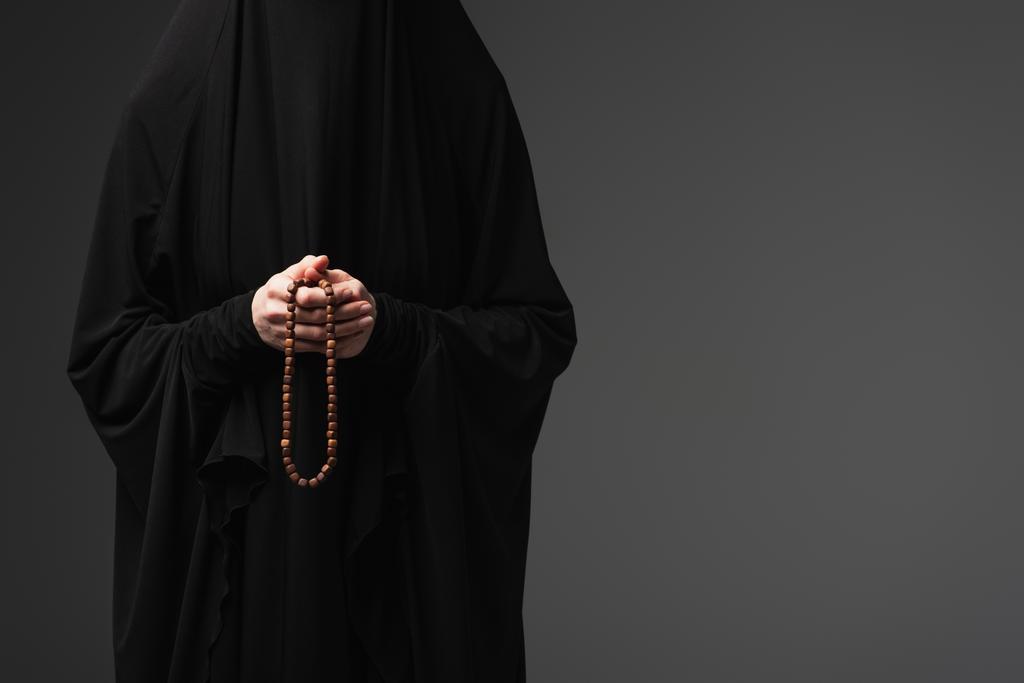 Teilansicht einer muslimischen Nonne in schwarzer Abaya, die Gebetsperlen isoliert auf dunkelgrau hält - Foto, Bild