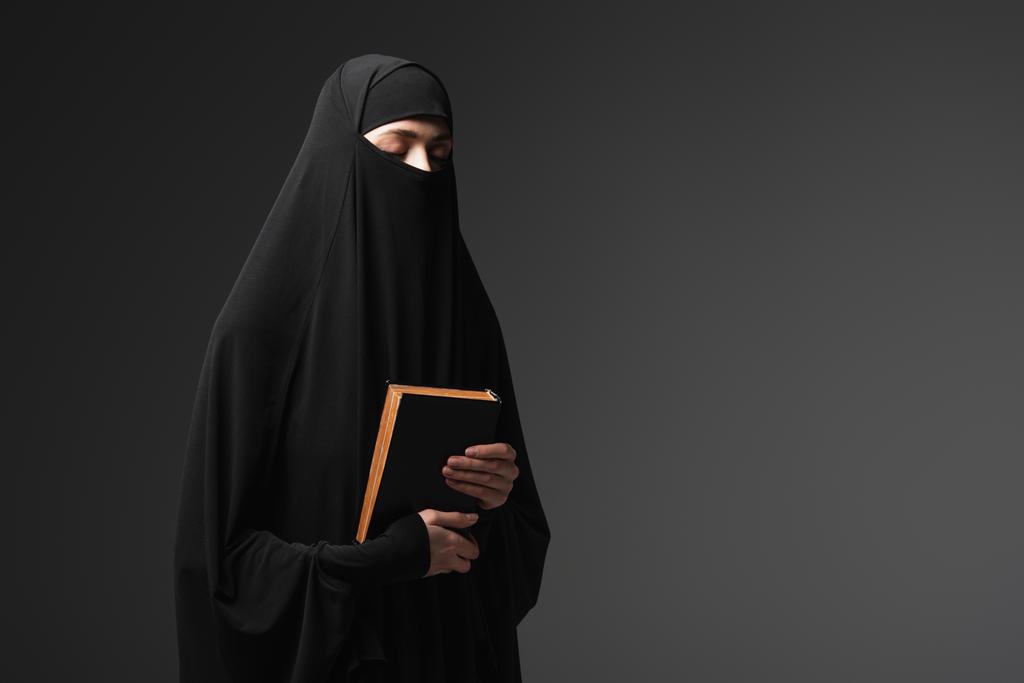 młody muzułmańska zakonnica gospodarstwa koran podczas modlitwy z zamkniętymi oczami odizolowany na czarny - Zdjęcie, obraz
