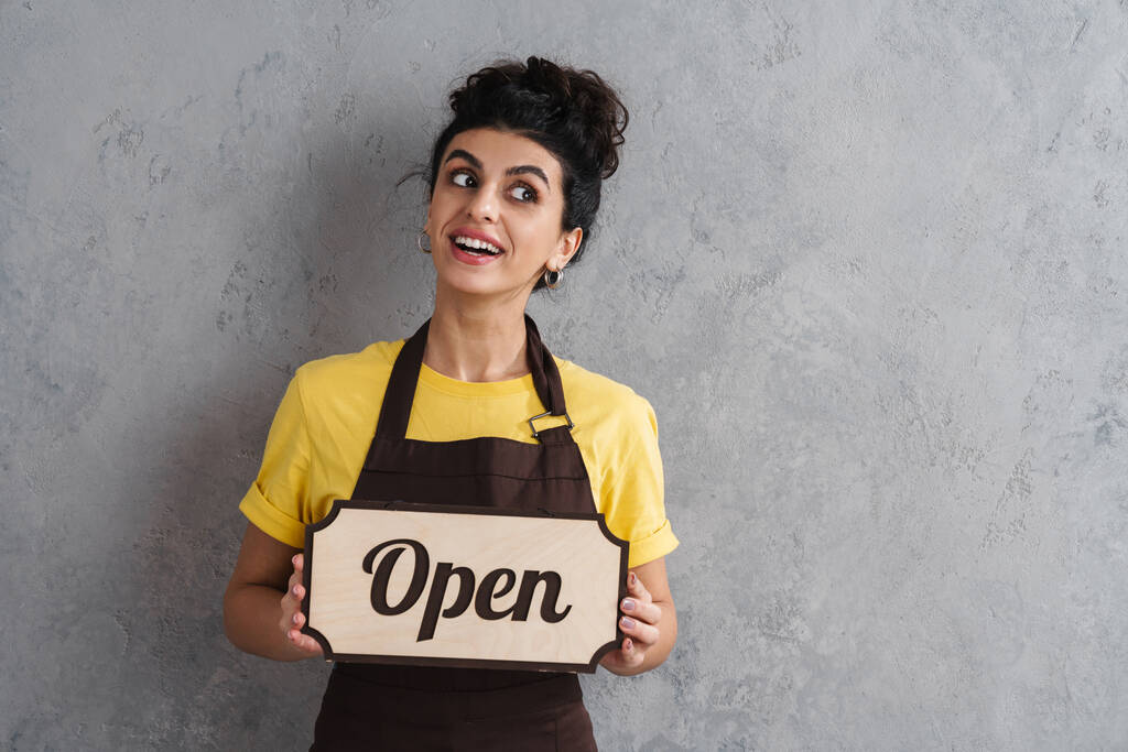 Joyful beautiful brunette girl in apron showing open sign board isolated over grey wall - Photo, Image