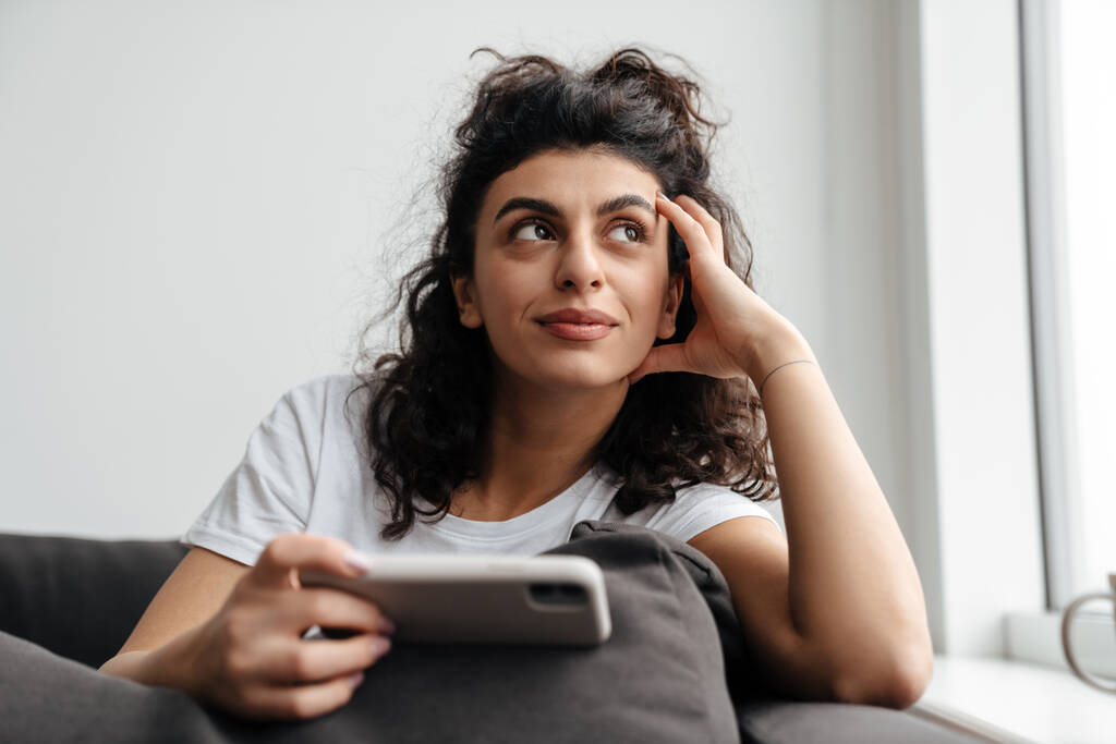 Удовлетворенная брюнетка молодая женщина с помощью мобильного телефона, сидя на диване дома - Фото, изображение