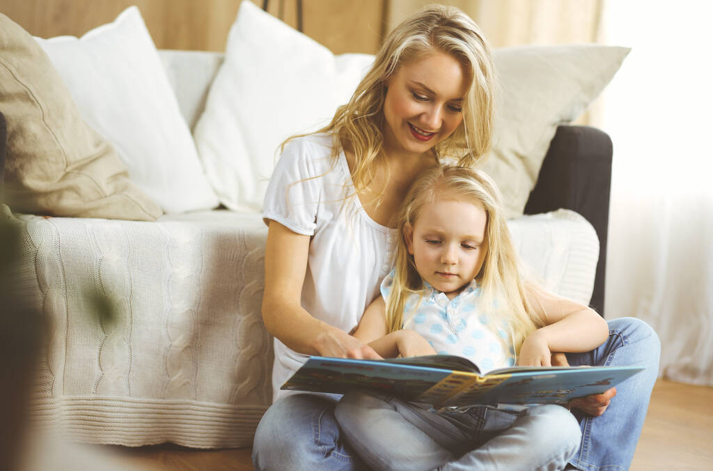 Happy family. Blonde young mother reading a book to her cute daughter while sitting at wooden floor. Motherhood concept - Photo, Image