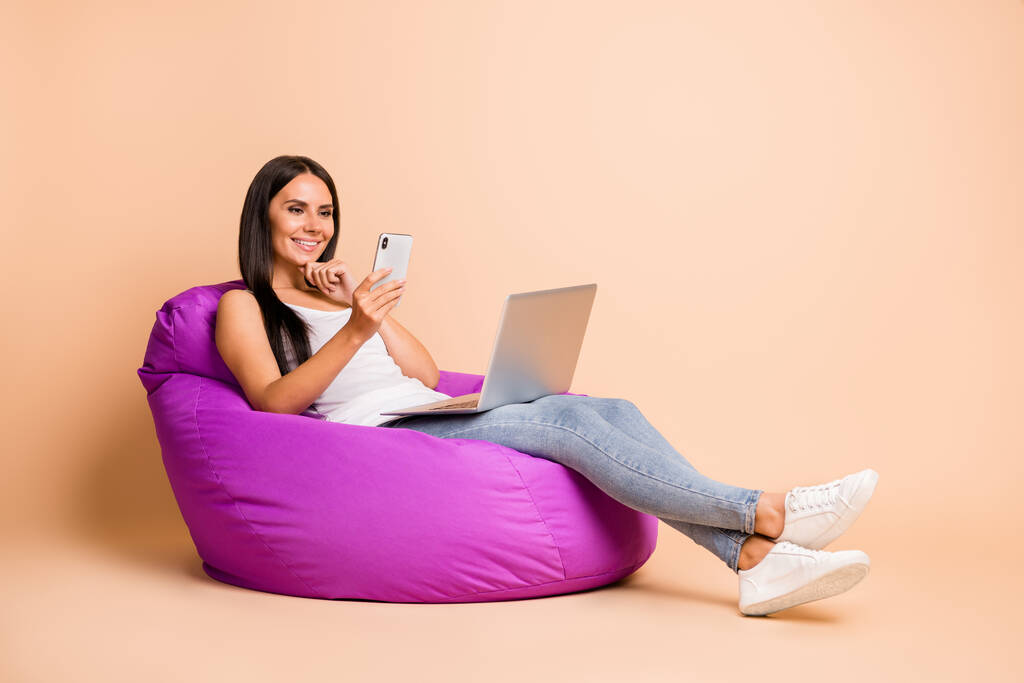 Foto Porträt Ganzkörperansicht der Frau mit Laptop-SMS am Telefon sitzen in Sitzsack Stuhl isoliert auf pastellbeige farbigem Hintergrund - Foto, Bild