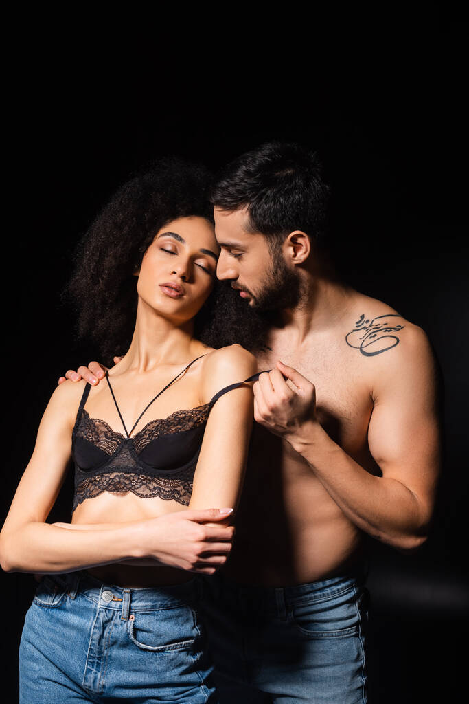 Shirtless man taking off lace bra from african american woman with closed eyes isolated on black  - Photo, Image