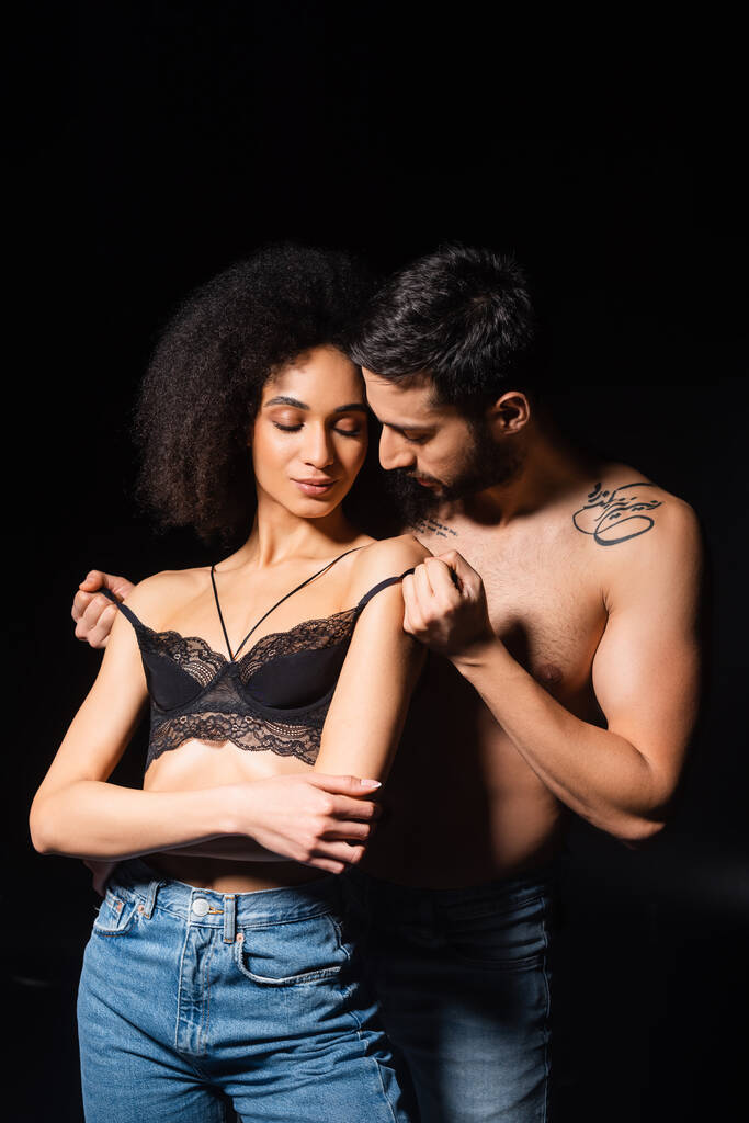 Man undressing seductive african american girlfriend in bra isolated on black  - Photo, Image