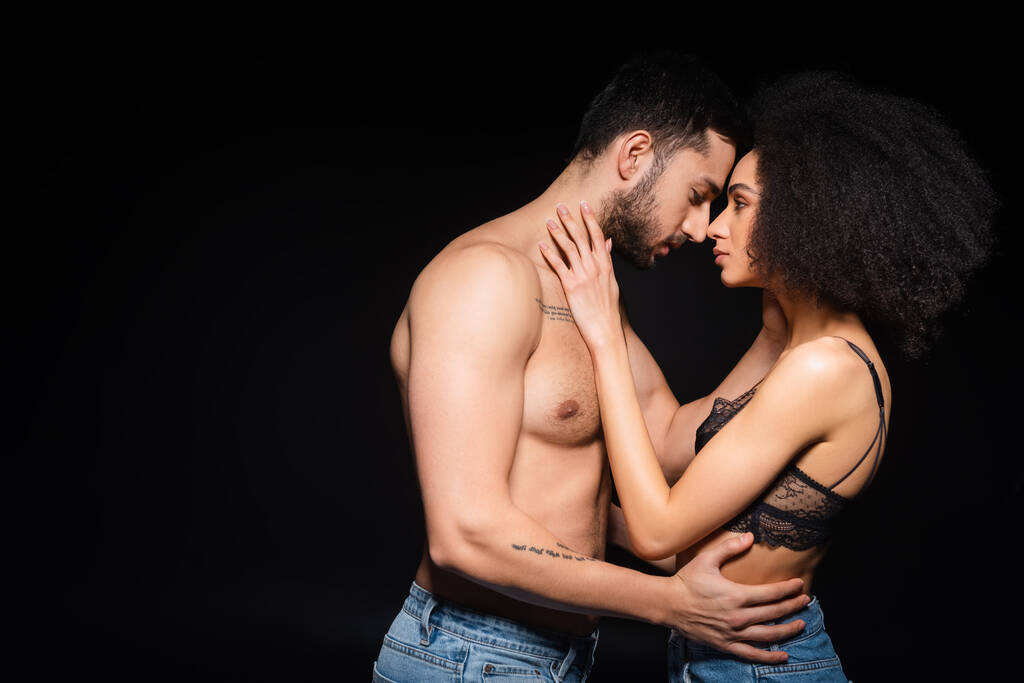 Vista lateral del hombre tatuado abrazando a la sexy mujer afroamericana aislada en negro  - Foto, imagen