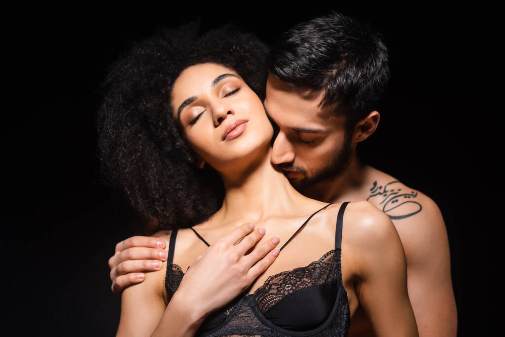 Татуированный мужчина целует шею великолепной африканской девушки в бюстгальтере, изолированном на черном  - Фото, изображение