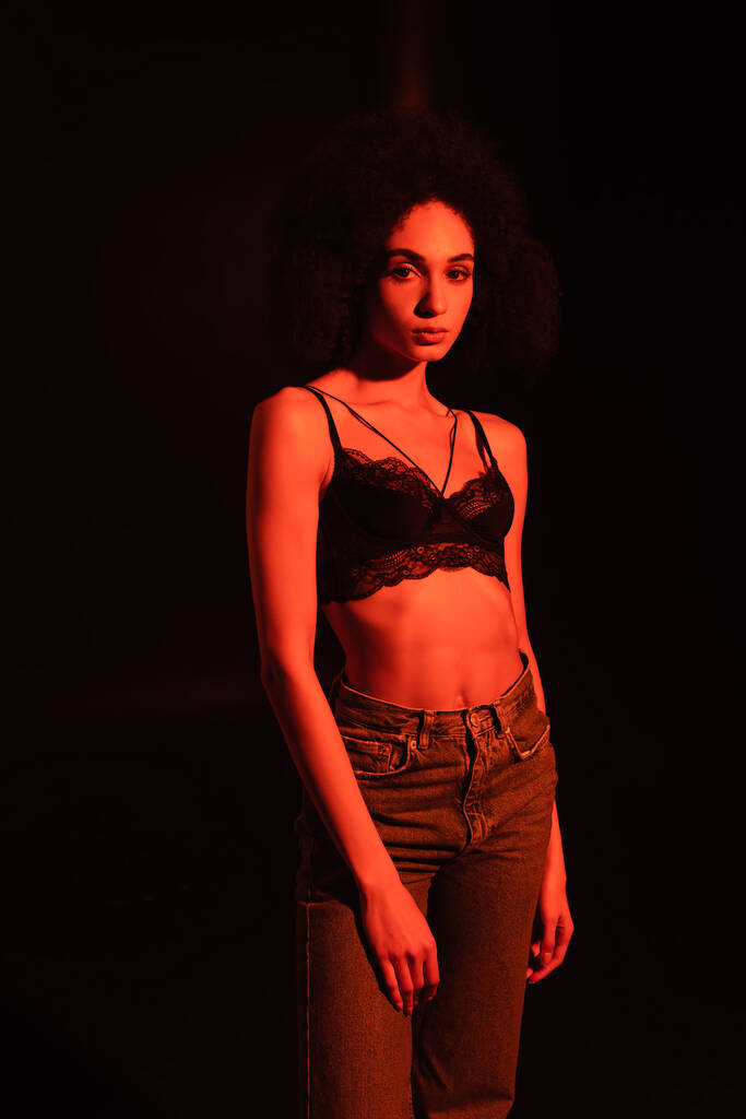 Young african american woman in jeans and bra looking at camera isolated on black with red lighting  - Photo, Image