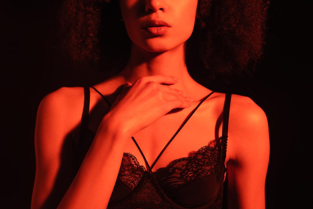 Vista ritagliata di donna afro-americana in reggiseno toccare il collo isolato su nero con luce rossa  - Foto, immagini