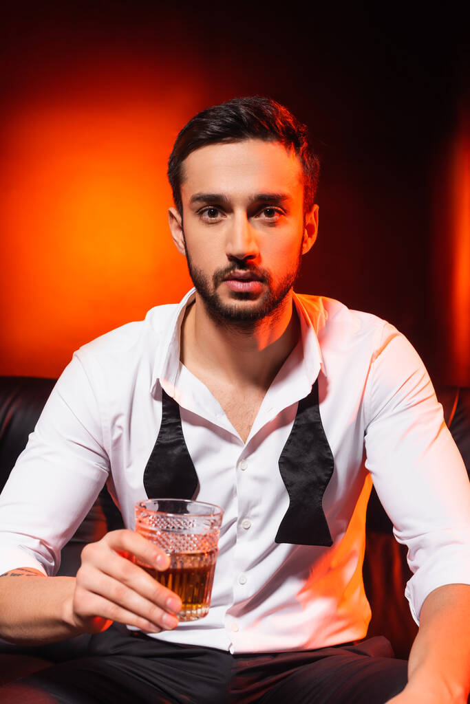 Mężczyzna w formalnym ubraniu trzyma szklankę whisky na kanapie na czarnym tle z czerwonym oświetleniem - Zdjęcie, obraz