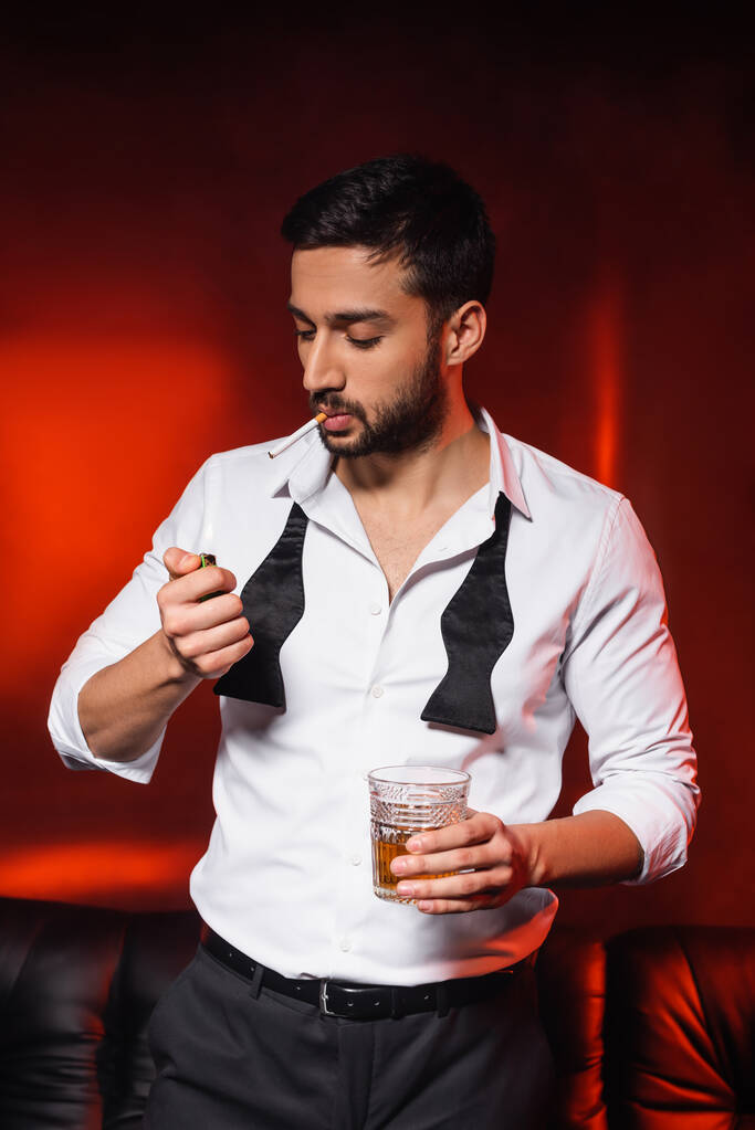 Hombre barbudo con cigarrillo y whisky sosteniendo encendedor sobre fondo negro con iluminación roja - Foto, Imagen