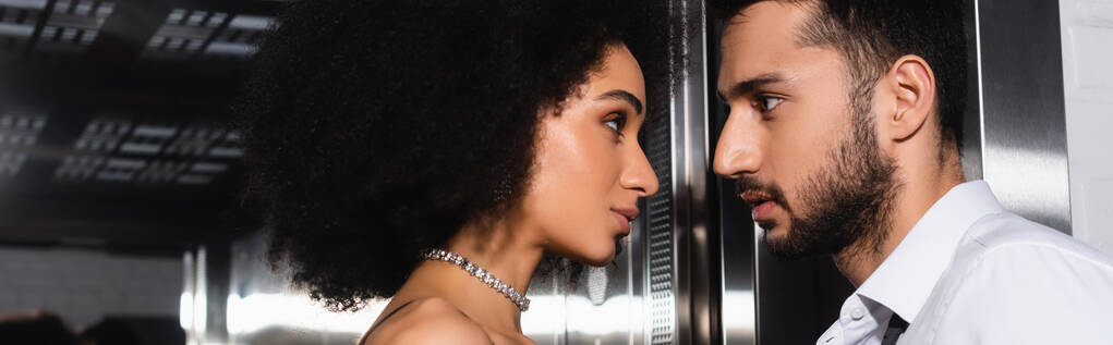 Vista lateral de mujer afroamericana en collar mirando novio cerca del ascensor, pancarta  - Foto, imagen
