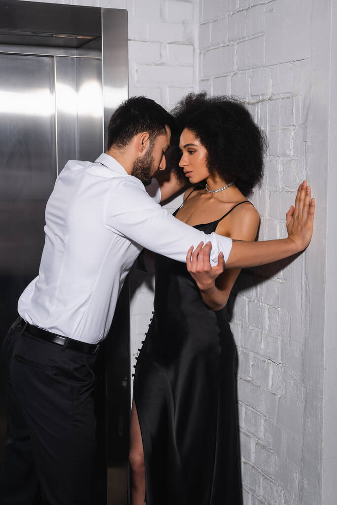 Hombre de pie cerca de la novia afroamericana en vestido cerca del ascensor  - Foto, imagen