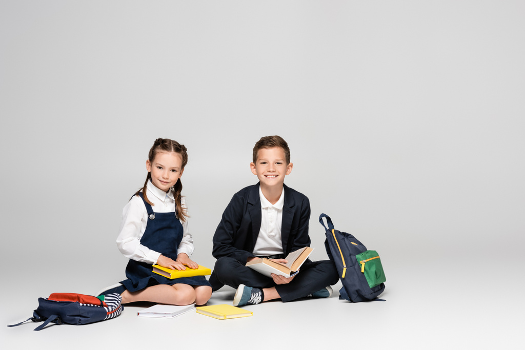 heureux écoliers en uniforme prestigieux assis avec des sacs à dos et des livres sur gris - Photo, image