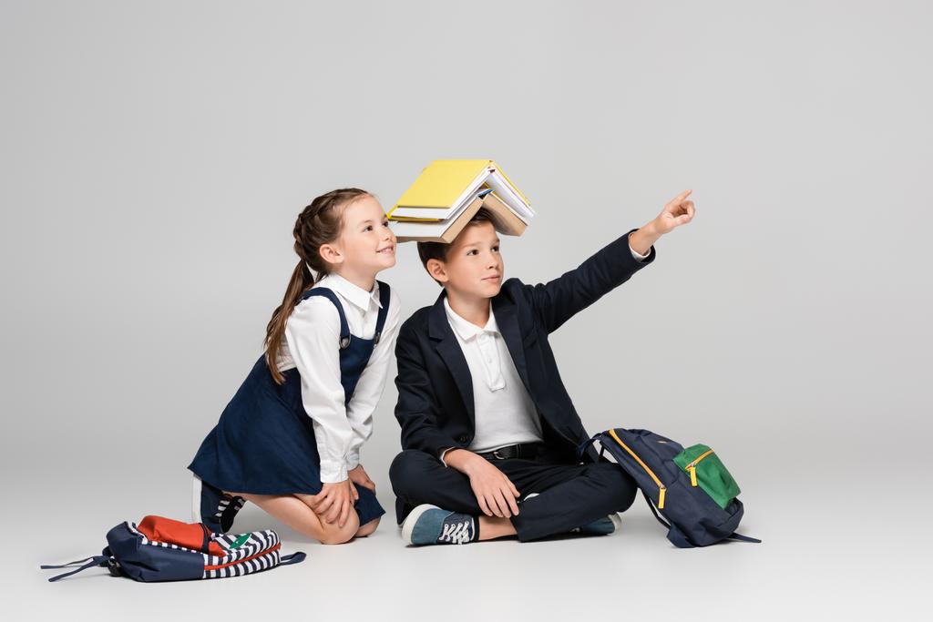 uczeń z książkami na głowie wskazując palcem w pobliżu wesołej dziewczyny na szarości - Zdjęcie, obraz