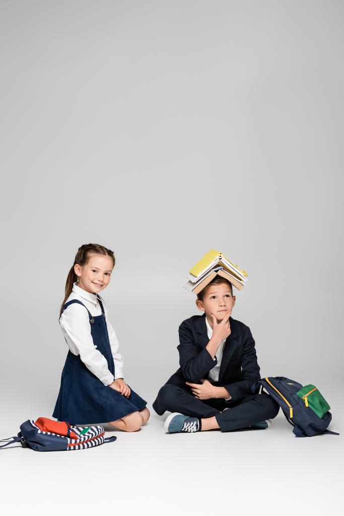 pensive schooljongen met boeken op hoofd zitten in de buurt vrolijk meisje op grijs - Foto, afbeelding