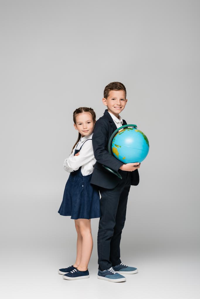felice scolaro tenendo il globo mentre in piedi con la ragazza in abito grigio - Foto, immagini