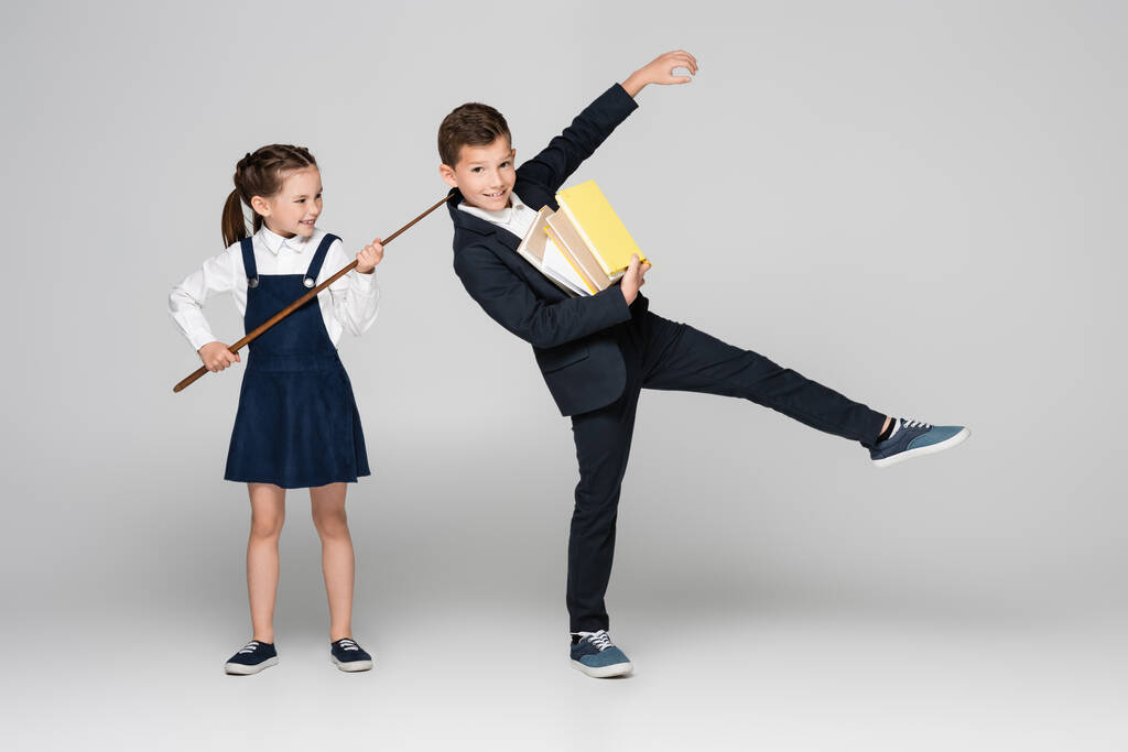 scolaretta tenendo puntare bastone vicino allegro scolaro in uniforme tenuta mucchio di libri su grigio - Foto, immagini