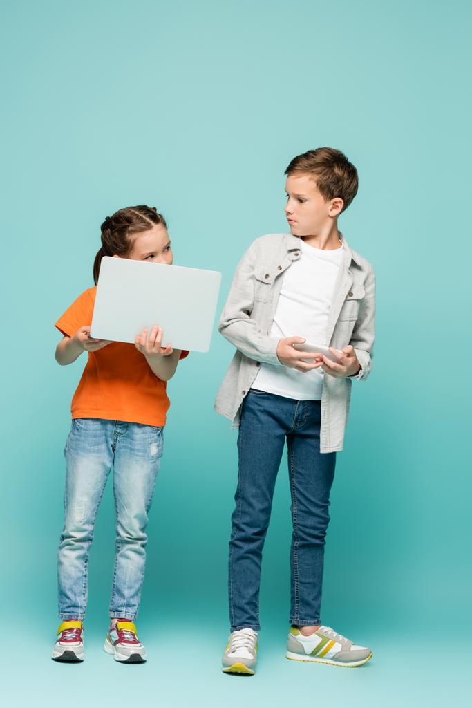 meisje met laptop kijken naar jongen holding smartphone op blauw - Foto, afbeelding