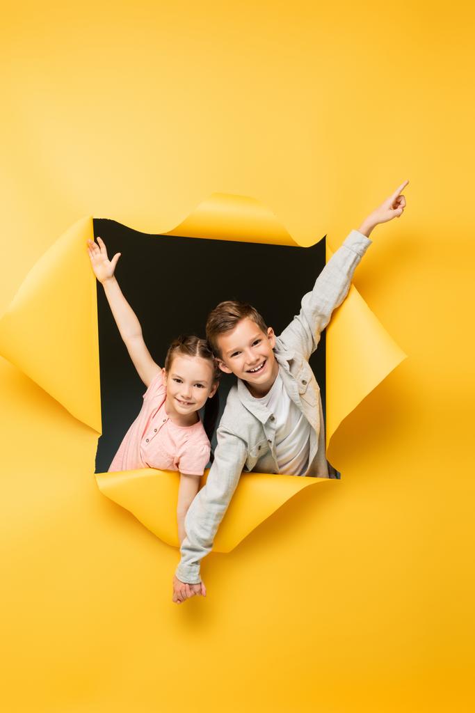 niños felices tomados de la mano y sonriendo a través del agujero en el fondo amarillo - Foto, imagen