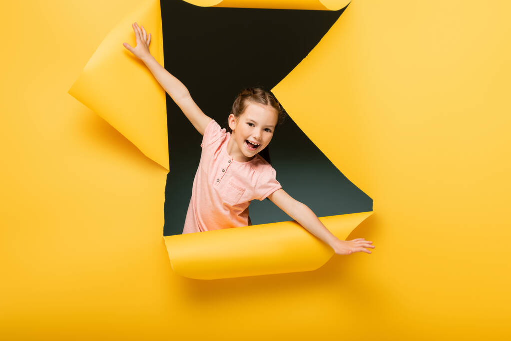 ενθουσιασμένο παιδί με απλωμένα χέρια κοιτάζοντας κάμερα μέσα από σχισμένη τρύπα στο κίτρινο φόντο - Φωτογραφία, εικόνα