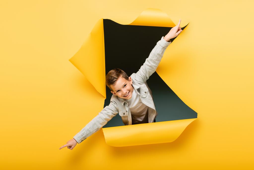 радостный мальчик с протянутыми руками, смотрящий в камеру через дыру на желтом фоне - Фото, изображение
