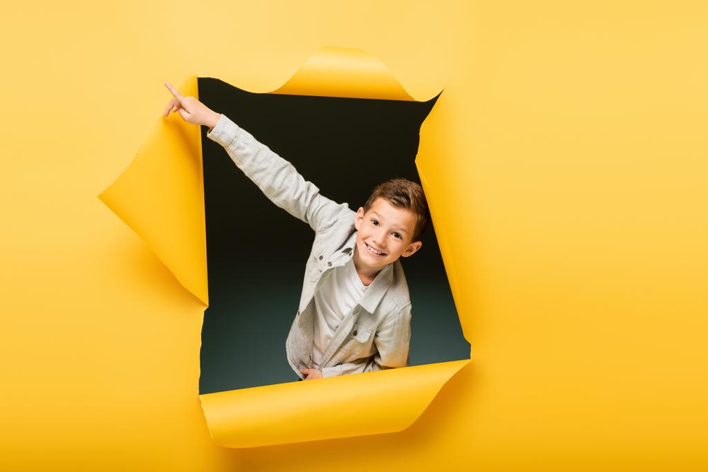 šťastný chlapec ukazuje prstem a dívá se na kameru přes roztrženou díru na žlutém pozadí - Fotografie, Obrázek