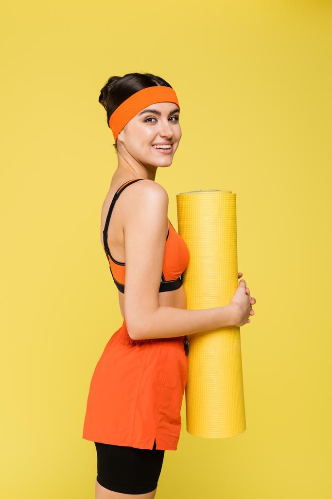 smiling sportswoman com tapete de fitness olhando para a câmera isolada no amarelo - Foto, Imagem