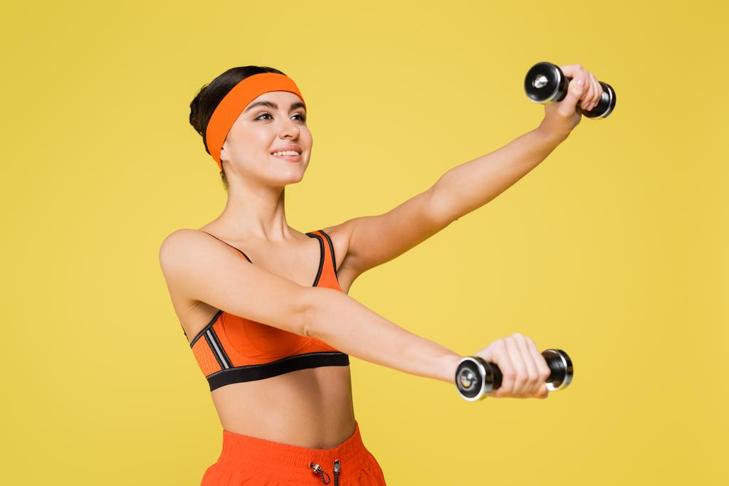 黄色に隔離されたダンベルとオレンジスポーツウェアのトレーニングで笑顔のスポーツ女性 - 写真・画像